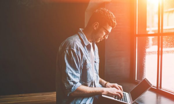 Caucasian male freelancer sitting near cafeteria window and typing new article in modern laptop application, young man blogger browsing wireless internet and updating profile in social networks